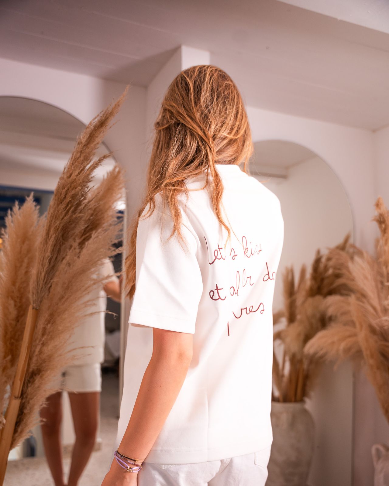 A woman viewed from behind wearing a white t-shirt with cursive French text on the back, standing in a bright interior with pampas grass decor.