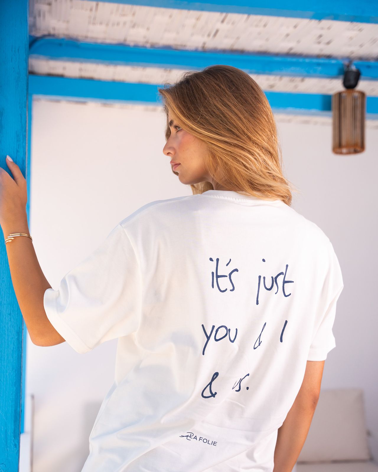 A woman wearing a white oversized t-shirt with 'it's just you & I & us.' printed in navy blue script on the back, shown from behind in a brightly lit setting with a blue and white wall.