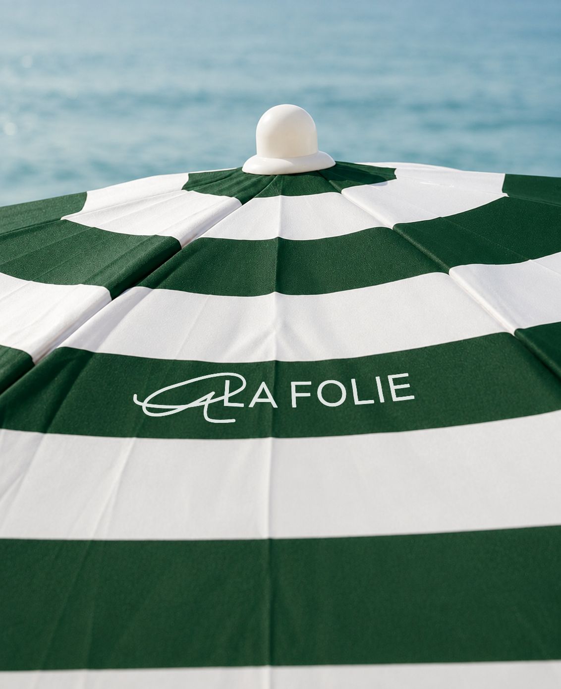 A close-up of a green and white striped beach umbrella with the brand name 'LA FOLIE' printed in white on a green stripe, with a blurred blue ocean background.