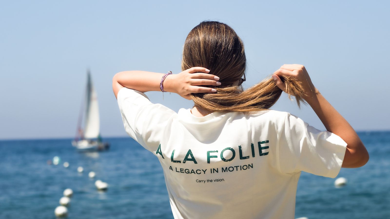 A woman seen from behind wearing a white graphic T-shirt with 'A LA FOLIE A LEGACY IN MOTION' printed on the back, standing by the ocean with a sailboat in the background.