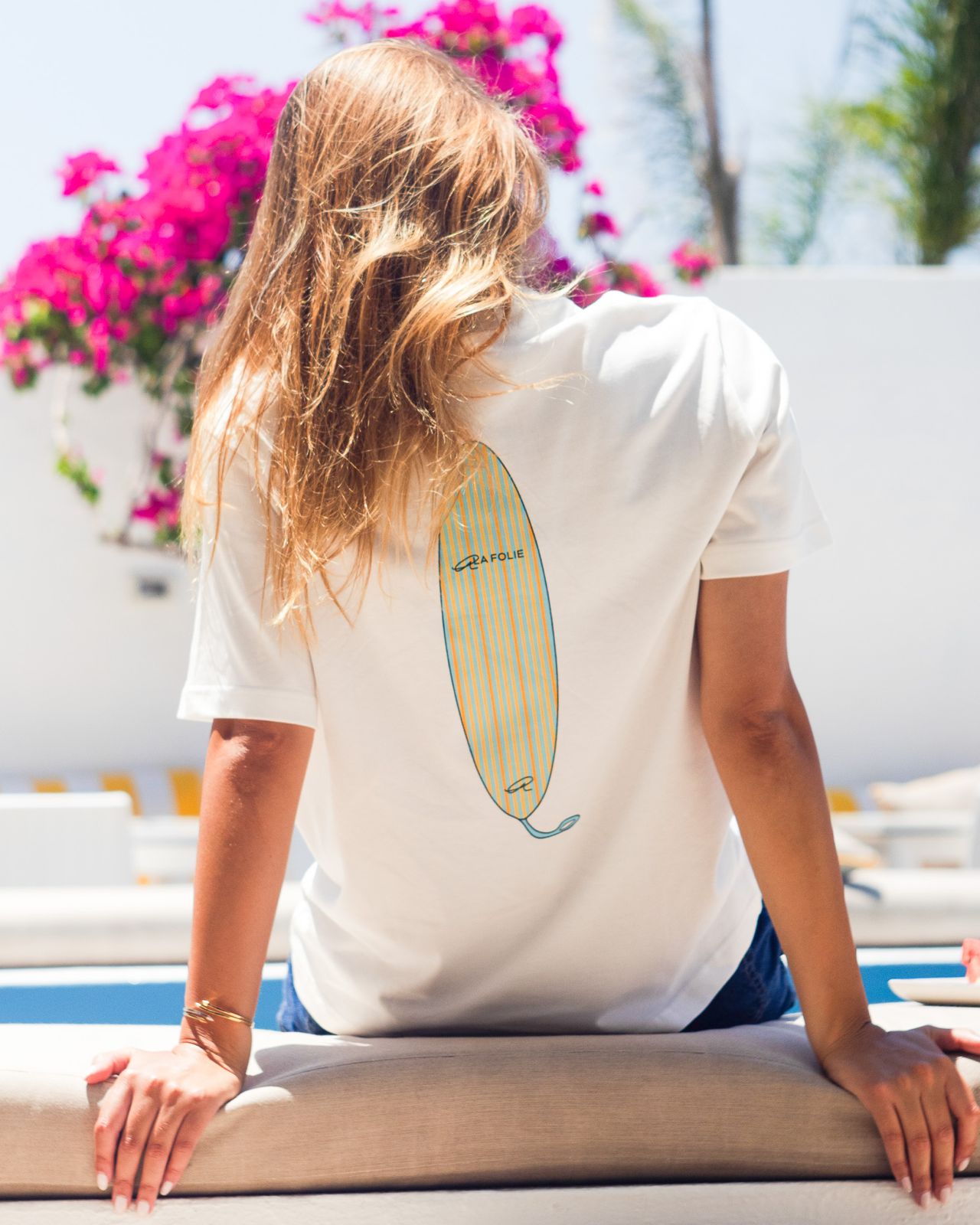 A woman with long blonde hair is seen from behind, wearing a white t-shirt with a yellow and blue surfboard graphic on the back. She is leaning on a beige lounge chair in an outdoor setting with bright pink bougainvillea flowers and a white wall in the background.